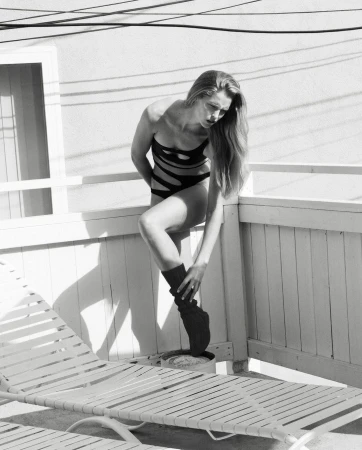 Monochrome portrait of a woman in a black swimsuit and leg warmer leaning against a white railing near a deck chair.