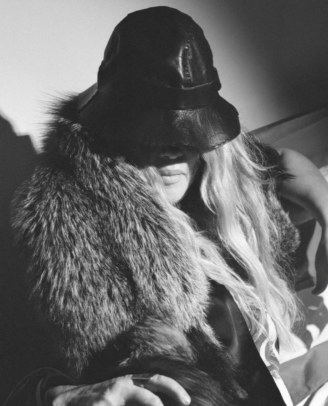 Black and white portrait of a woman wearing a leather hat and large fur collar, partially obscuring her face with hat shadow.