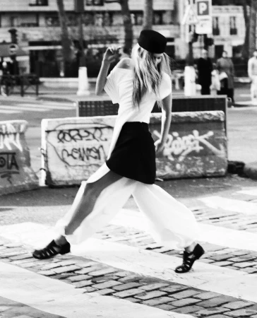 Black and white fashion shot of a woman in white pants and top with black hat crossing a street on a crosswalk with graffiti barriers.