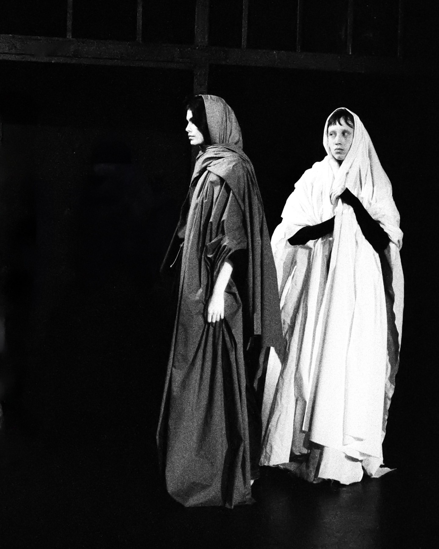 Two figures draped in white and gray robes stand in a dark, shadowy indoor setting, with light highlighting the fabric's texture.