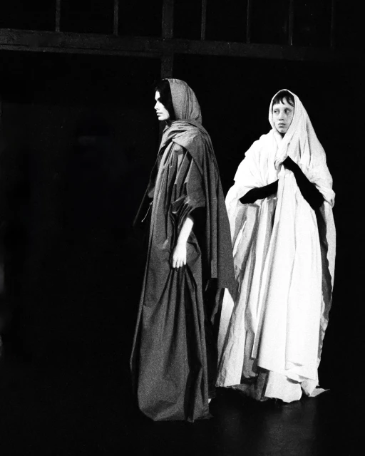 Two figures draped in white and gray robes stand in a dark, shadowy indoor setting, with light highlighting the fabric's texture.