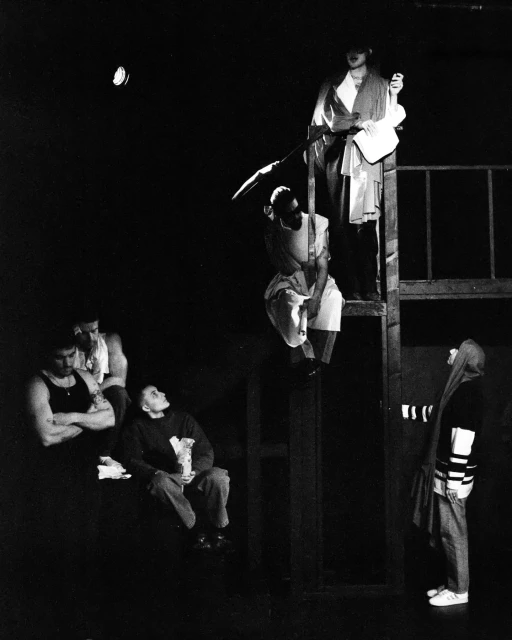 A group of people on and around a wooden platform under spotlighting, creating a dramatic, theatrical black-and-white scene.