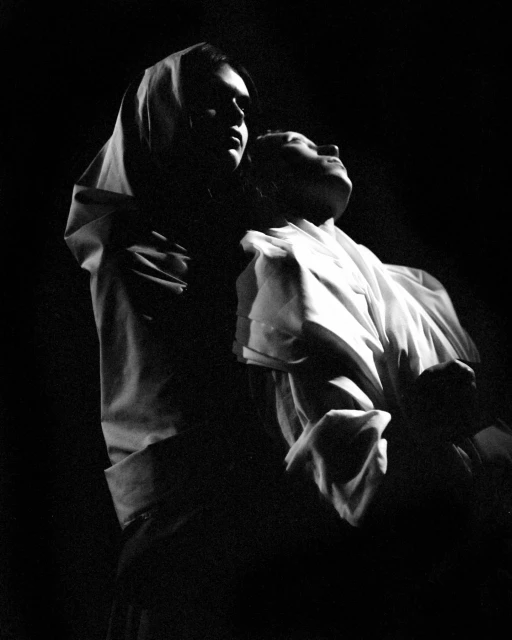 Black and white portrait of two women, one holding the other, wearing a headscarf and white clothing in a dark, indoor setting.