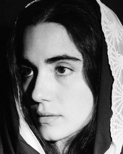 Close-up portrait of a young adult female with dark hair wearing a light colored headscarf looking at the camera against a dark backdrop.