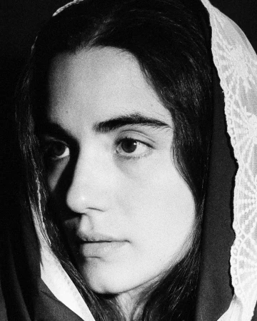 Close-up portrait of a young adult female with dark hair wearing a light colored headscarf looking at the camera against a dark backdrop.