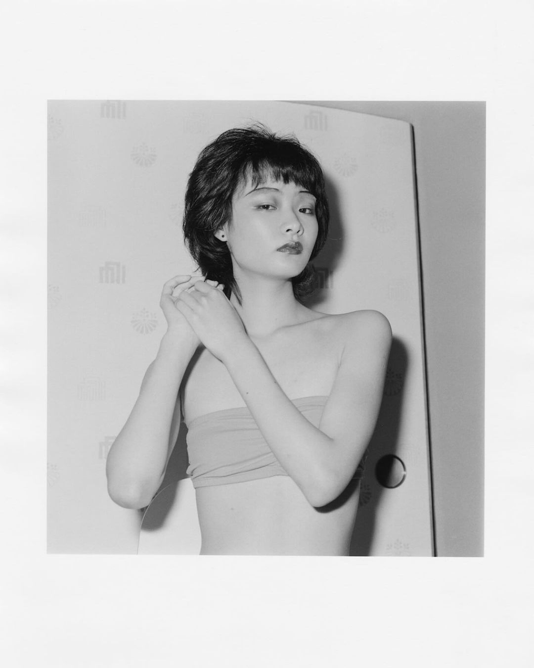 Monochrome portrait of an Asian woman with short dark hair, wearing a tube top, standing in front of a white panel in an indoor setting.