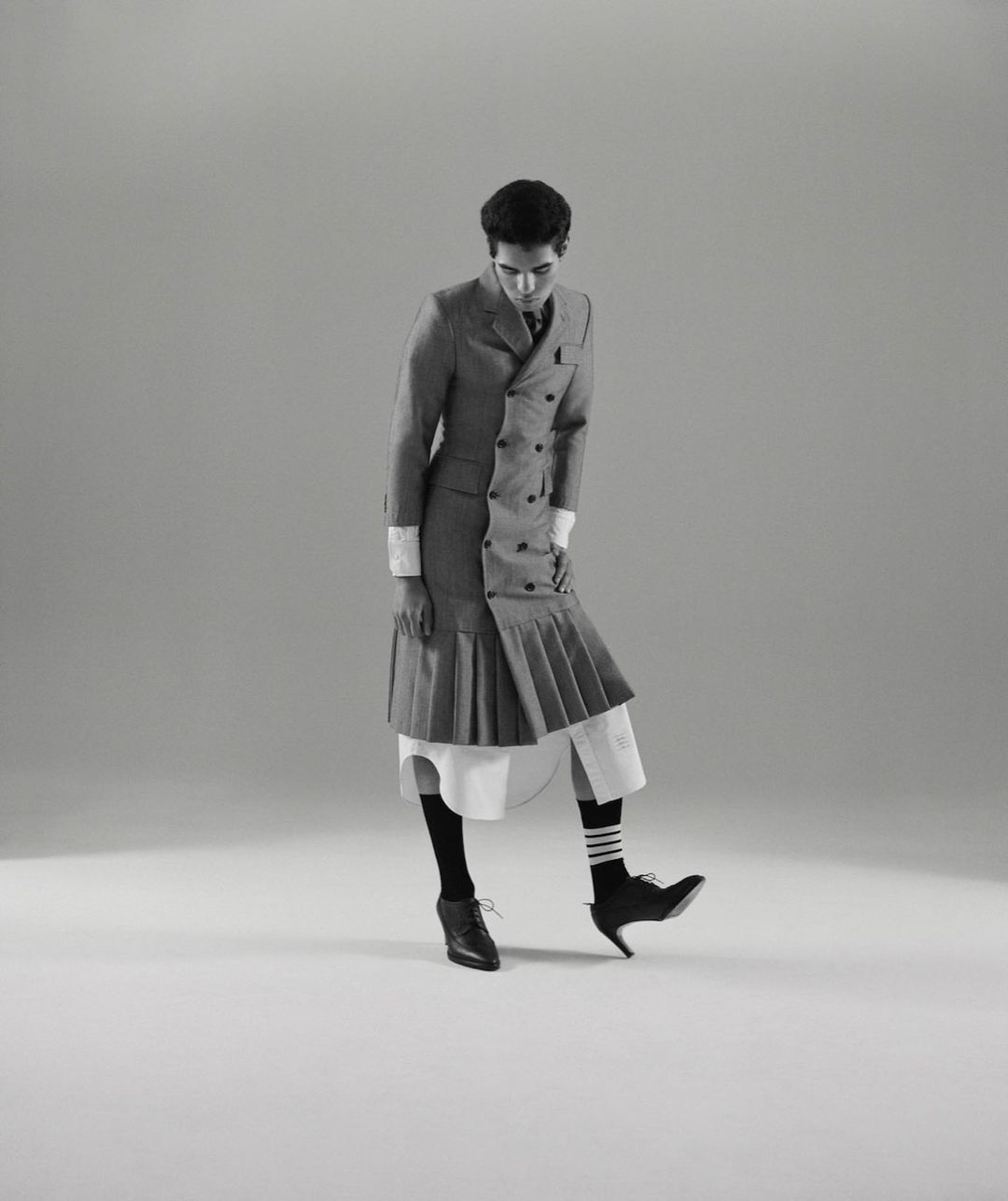 Person wearing a grey double-breasted coat with a pleated skirt and black high-heeled shoes stands in a neutral grey photo studio.