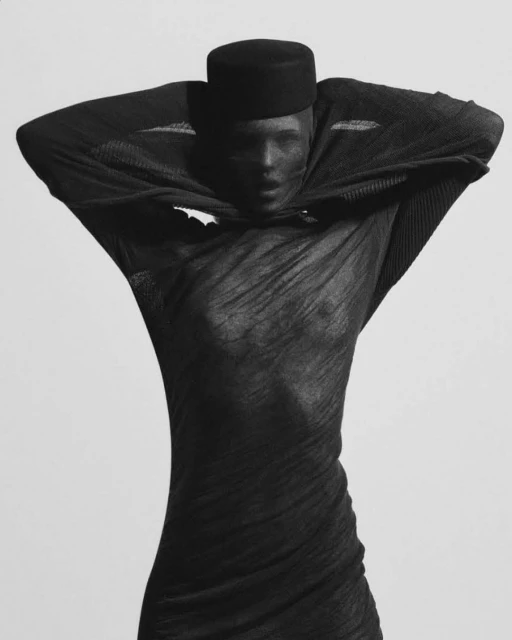 Monochrome portrait of a woman in a black sheer dress and a black cap, posing with her hands behind her head against a light background.