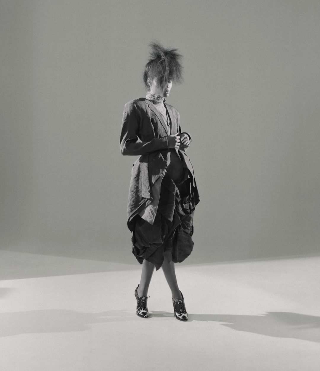 A fashion model in a jacket and textured skirt poses in a photo studio with bright lighting, creating a strong visual presence.