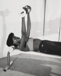 Black woman lying on a workout bench lifting chrome dumbbells in a London studio photographed by Danielle Neu.