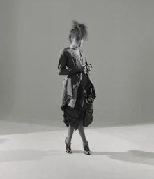 Model with voluminous spiky hair wearing a textured blazer and layered skirt against a grey background.