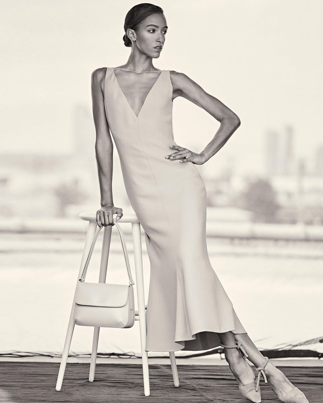 Elegant woman in a light-colored dress poses gracefully next to a stool and purse, set against a blurred cityscape backdrop.