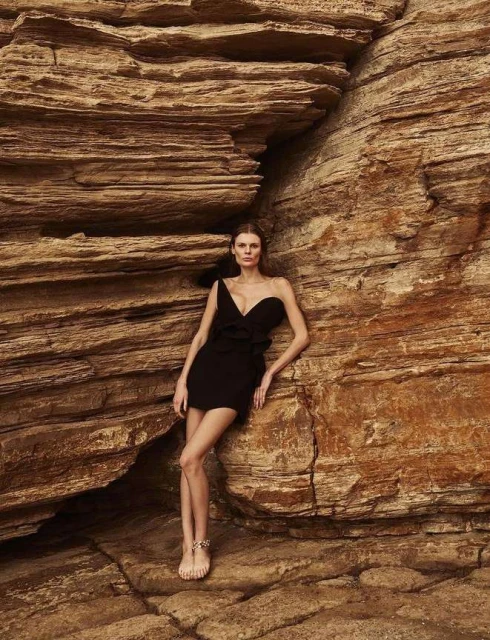 Woman in black mini dress poses barefoot against natural rock formation in outdoor fashion shot, medium shot, eye-level angle.