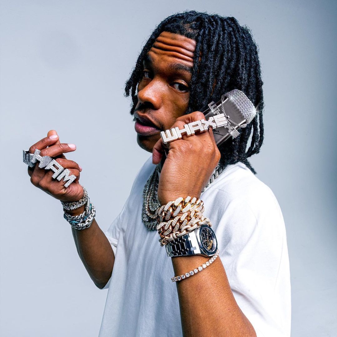 Close-up portrait of a young Black male musician wearing jewelry, with dreadlocks, posing with a microphone ring in a light studio setting.