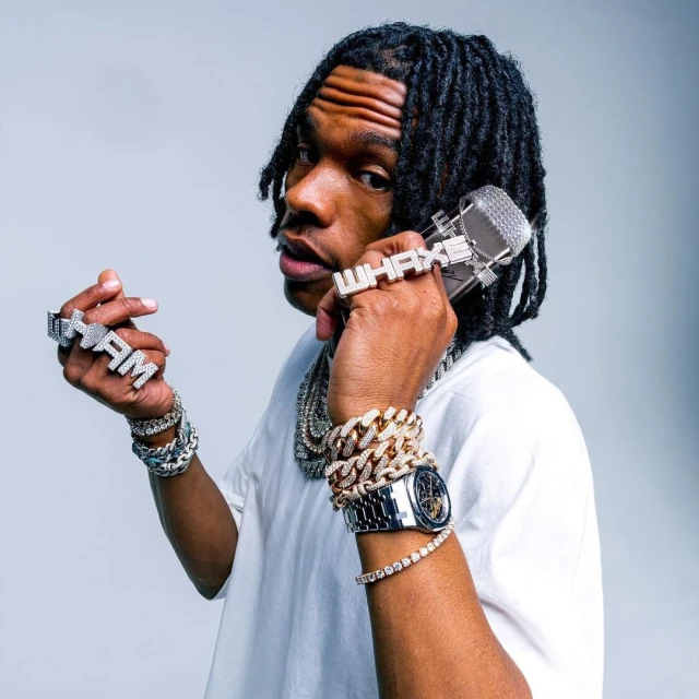 Close-up portrait of a young Black male musician wearing jewelry, with dreadlocks, posing with a microphone ring in a light studio setting.