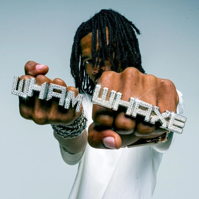 Close-up studio shot of a young adult male wearing jewelry, including letter rings with the words 'WHAM' and 'WHAXE'.