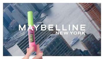 Close-up of a hand holding Maybelline mascara against a blurred cityscape background, featuring the Maybelline New York logo.