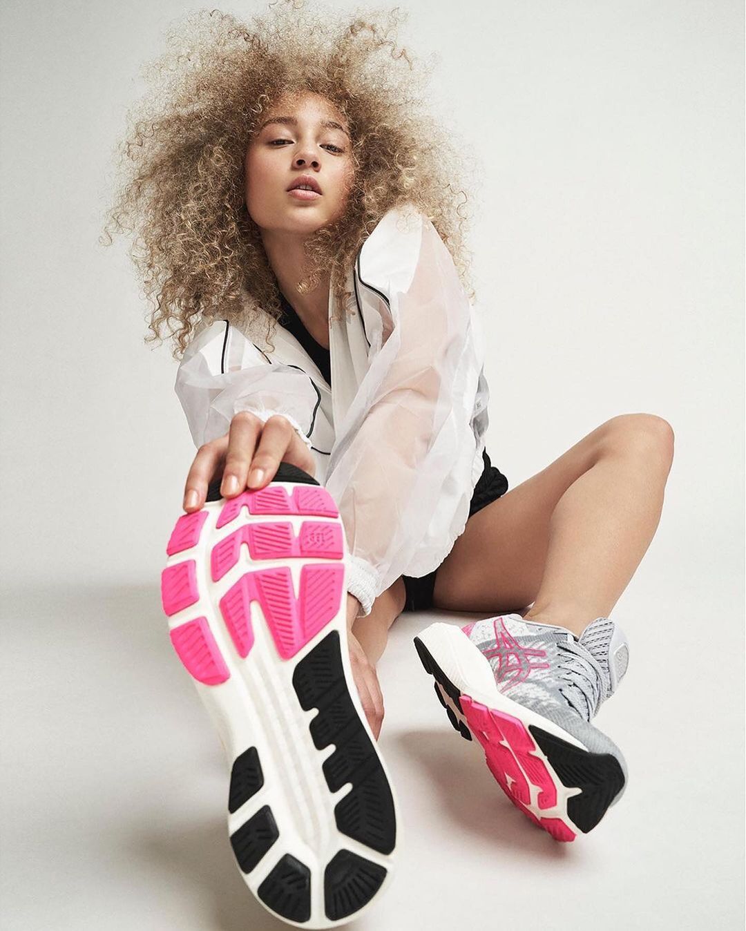 Athletic woman with curly hair is stretching her leg in front of a white background wearing a white jacket and black shorts, footwear closeup.