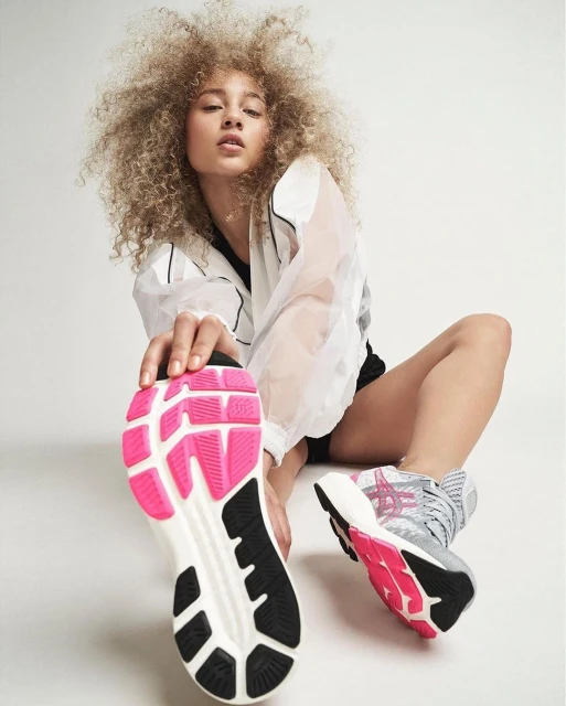 Athletic woman with curly hair is stretching her leg in front of a white background wearing a white jacket and black shorts, footwear closeup.
