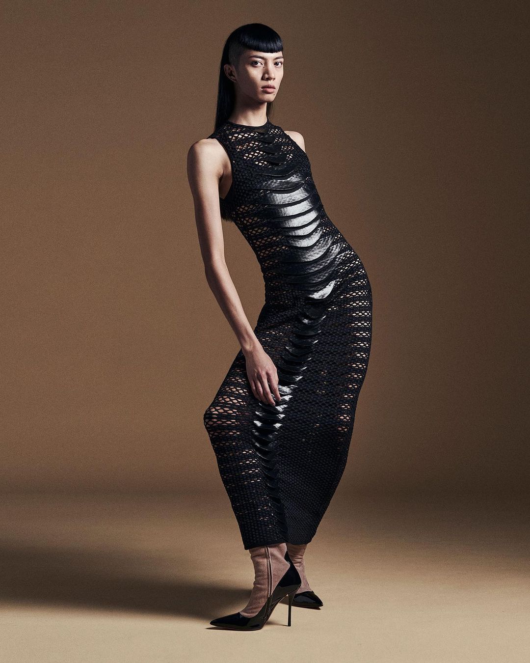 Fashion photo of a solo Asian female adult model in a black, textured dress and high heels, posing against a neutral backdrop.
