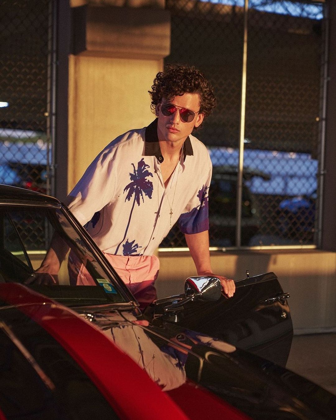 A curly-haired man wearing sunglasses leaning against a car door in an open-air garage by Danilo Hess