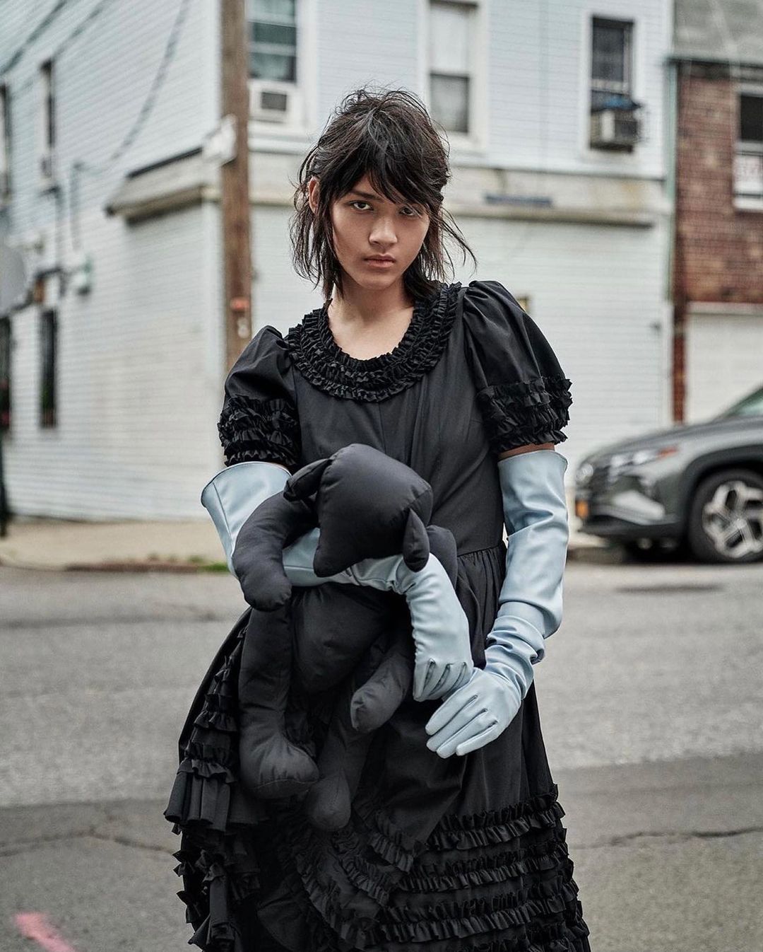 A person in a vintage-style black dress and gloves stands on a street holding a stuffed animal, with houses and a car in the background.