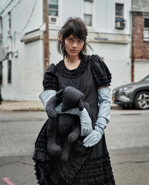 A person in a vintage-style black dress and gloves stands on a street holding a stuffed animal, with houses and a car in the background.