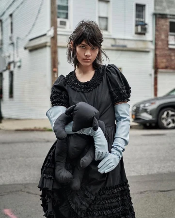 A person in a vintage-style black dress and gloves stands on a street holding a stuffed animal, with houses and a car in the background.