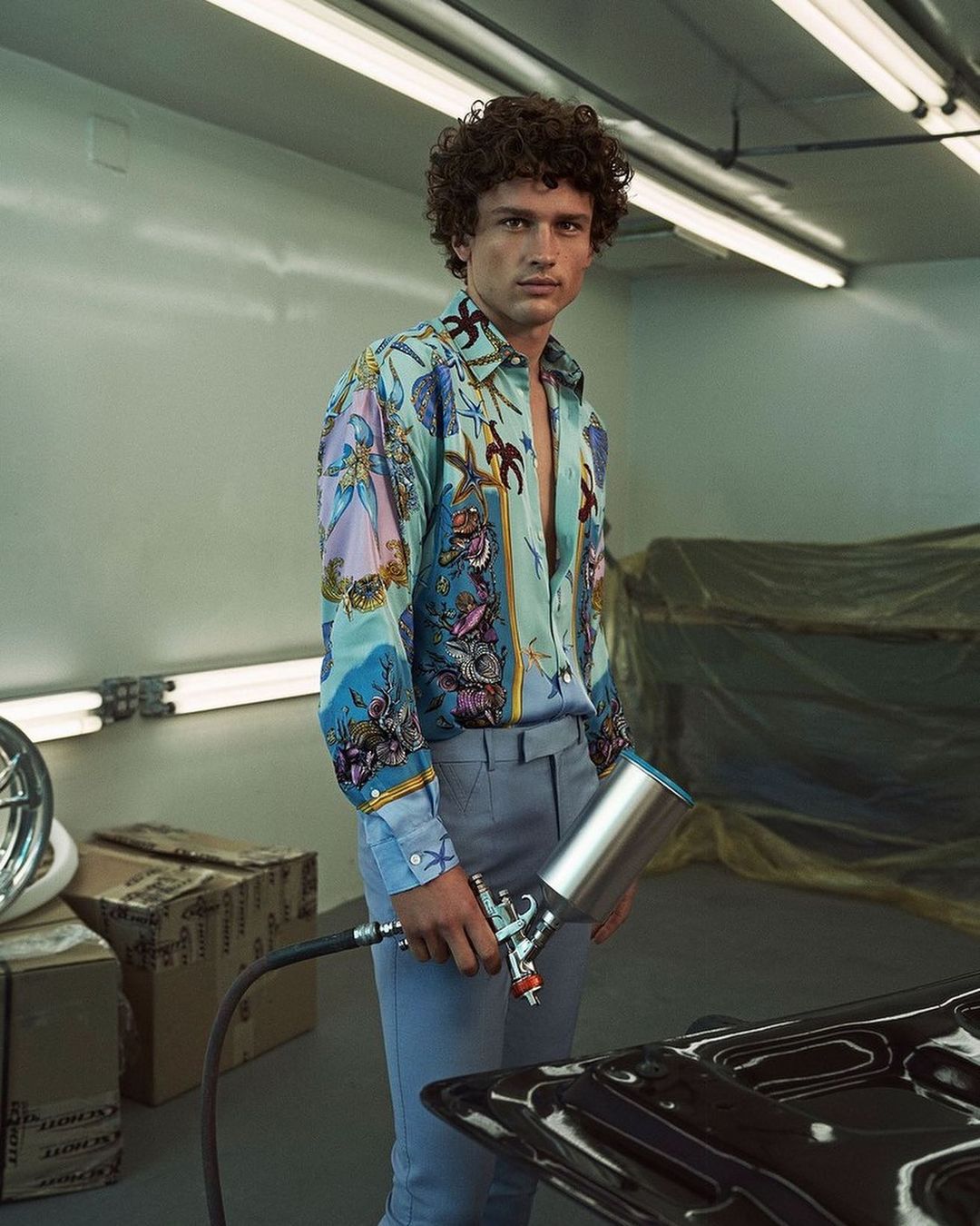 An adult man with curly brown hair, wearing a patterned shirt and grey pants, poses while holding a paint sprayer indoors.
