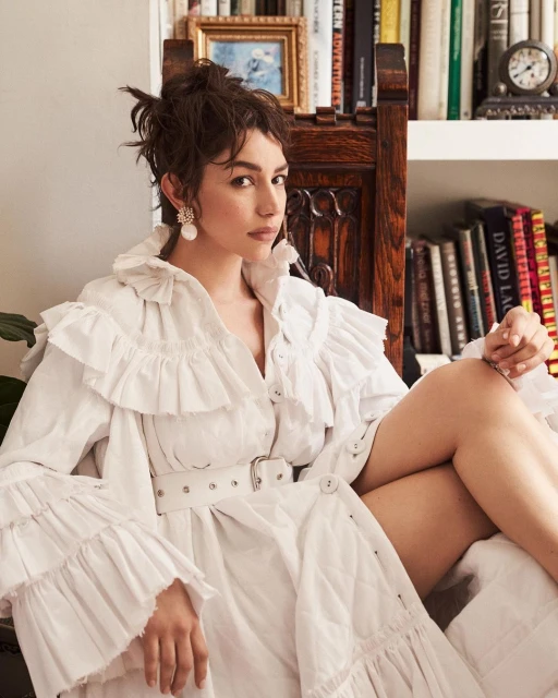 Woman with wavy brown hair poses wearing a white ruffled dress and belt indoors by books and a wood chair in soft light.