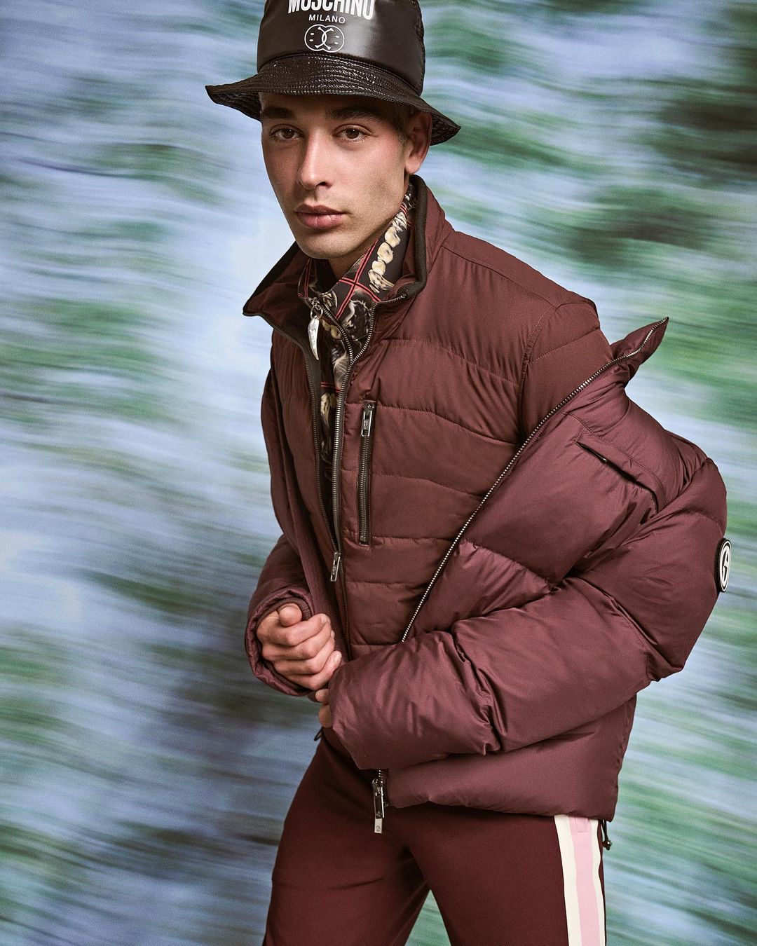 Young adult male in a burgundy puffer jacket and bucket hat poses against a blurred blue and green background in a fashion editorial shot.