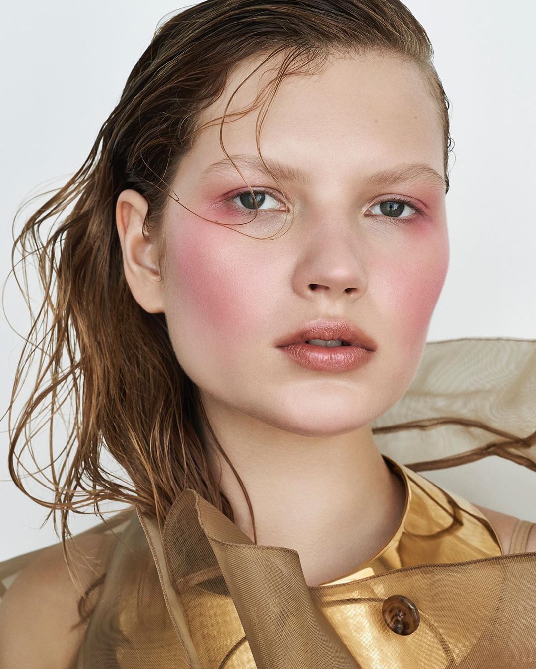 Close-up portrait of a young woman with wet brown hair, wearing a tan organza top with blush makeup, set against a white backdrop.