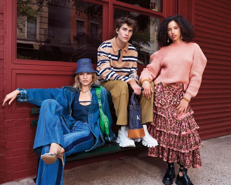 Three models wearing stylish clothing sit outside a brick building with window. One woman wears denim, another a pink sweater, the man a striped top.