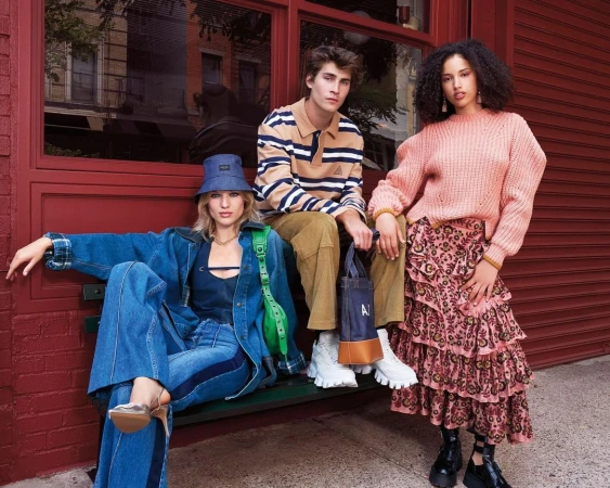 Three models wearing stylish clothing sit outside a brick building with window. One woman wears denim, another a pink sweater, the man a striped top.