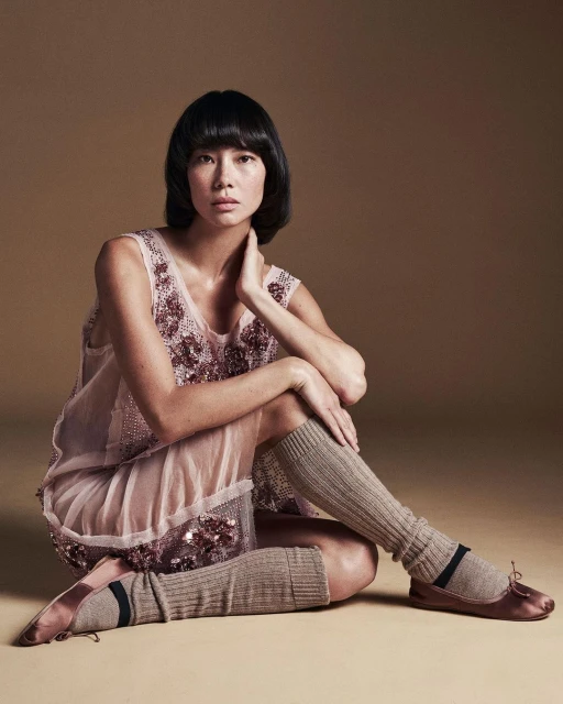 Portrait of an Asian woman with dark hair, wearing a light pink dress with sequin details, seated in a photo studio with a tan backdrop.