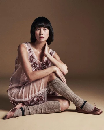 Portrait of an Asian woman with dark hair, wearing a light pink dress with sequin details, seated in a photo studio with a tan backdrop.