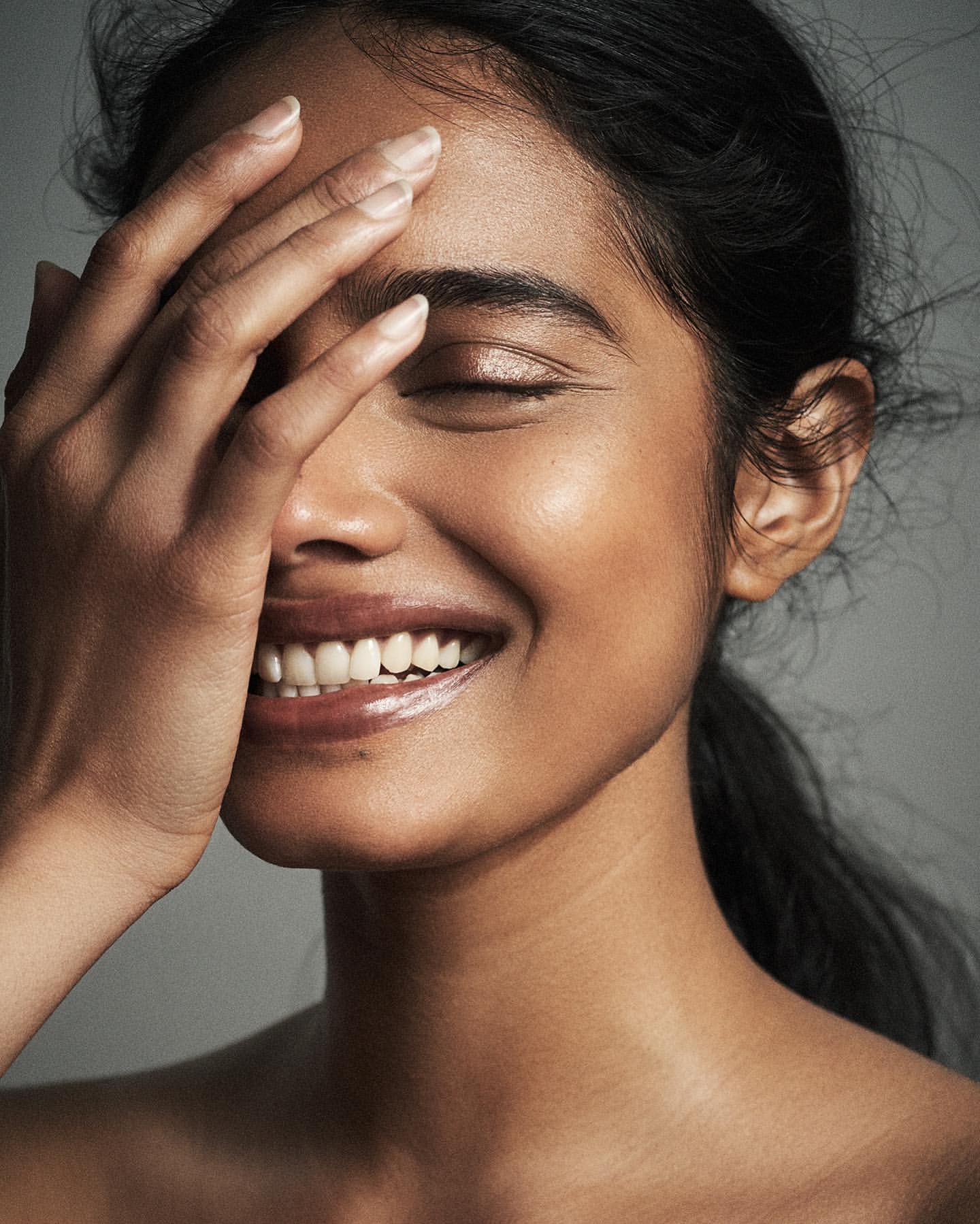 Close-up portrait of a smiling woman with hand raised to her forehead against a grey background by Danilo Hess