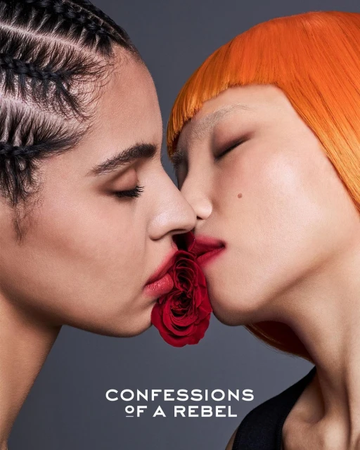 Two adult females in a close-up beauty shot, share a single red rose with their lips. The background is a simple gray color.