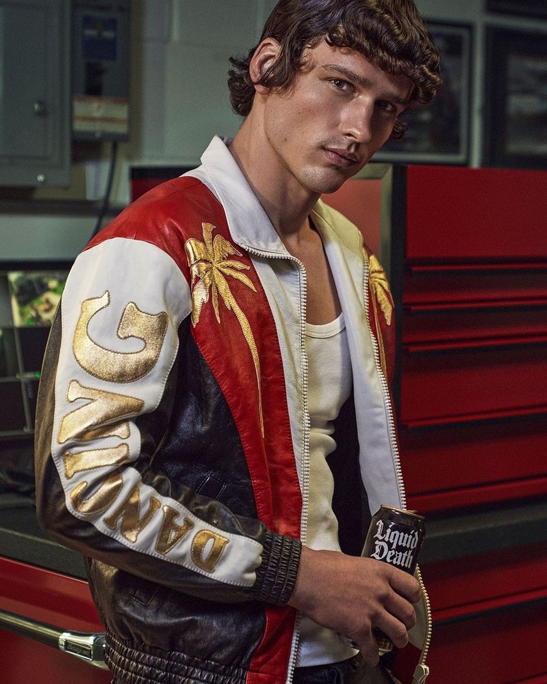 Man in a retro-style jacket posing indoors, holding a can of Liquid Death. The shot features a mix of white, red, and black colors.