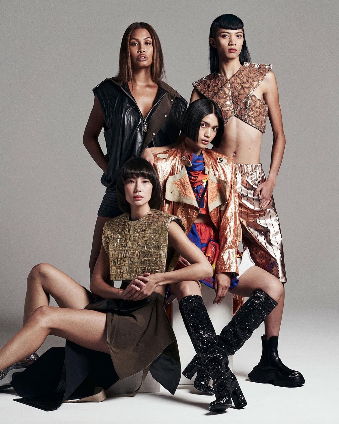 Four female fashion models in a group shot, wearing metallic modern-style clothing, captured in a studio setting against a grey background.