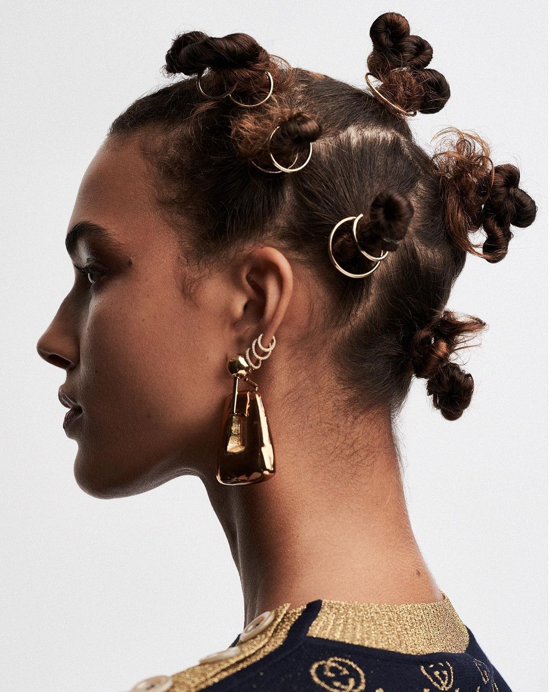 A side profile of a Black female model with dark brown hair styled in bantu knots and gold hoop earrings in a bright photo studio.