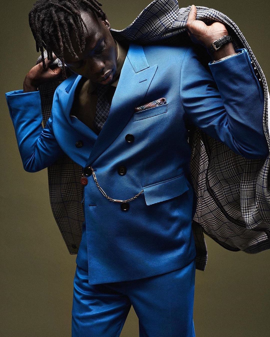 A man in a bright blue suit stands indoors, looking down and putting on a grey plaid jacket over his shoulders in a studio shot.