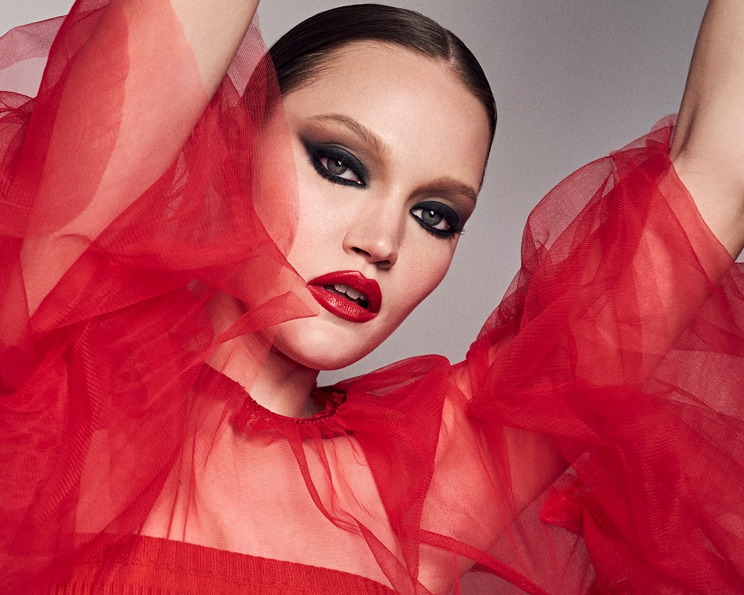 Close-up shot of a woman with dark eyeshadow and red lipstick wearing a red tulle top against a grey background in a studio setting.