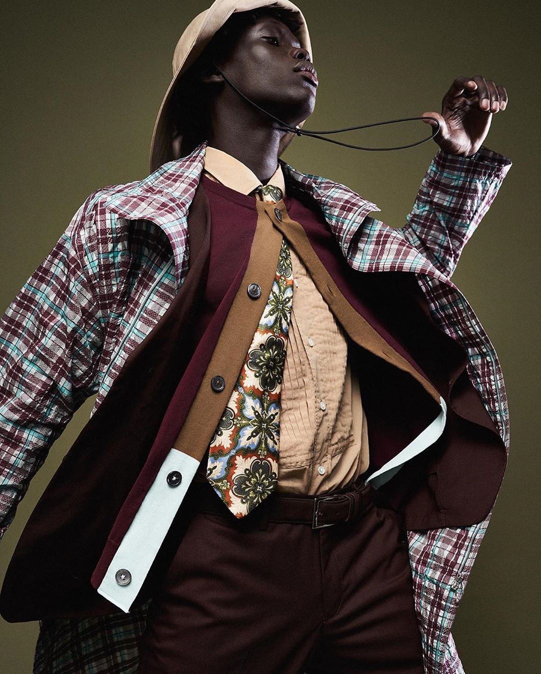 Dark-skinned adult man wearing a hat, patterned coat, vest, tie, and brown pants poses on a green background in a studio shot.