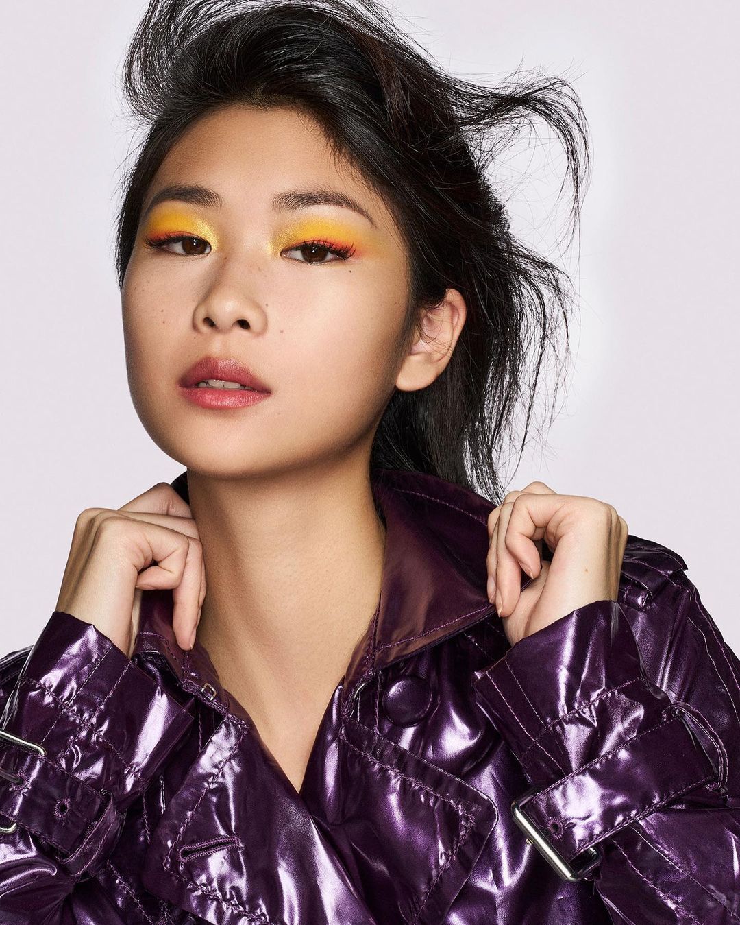 Close-up of a woman with vibrant yellow and orange eye makeup, wearing a shiny purple trench coat against a neutral background.