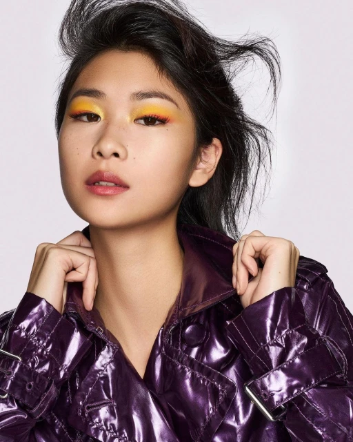 Close-up of a woman with vibrant yellow and orange eye makeup, wearing a shiny purple trench coat against a neutral background.