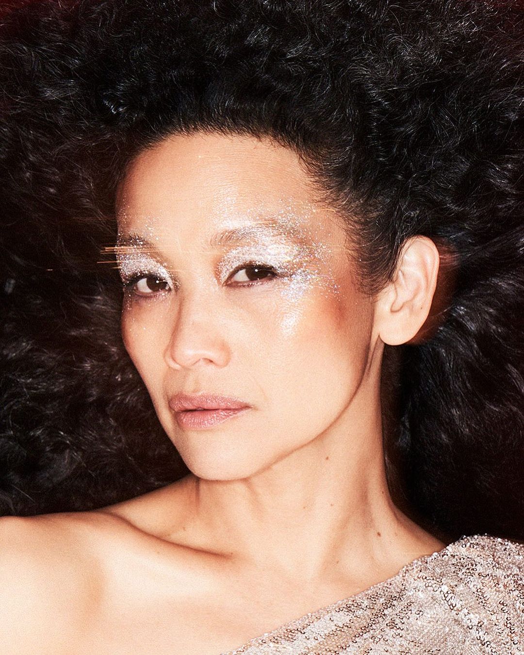 Close-up portrait of a woman with dark, curly hair and glittery eye makeup, wearing a sequined top against a dark background.