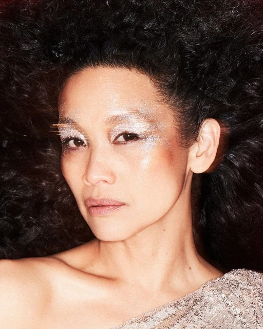 Close-up portrait of a woman with dark, curly hair and glittery eye makeup, wearing a sequined top against a dark background.