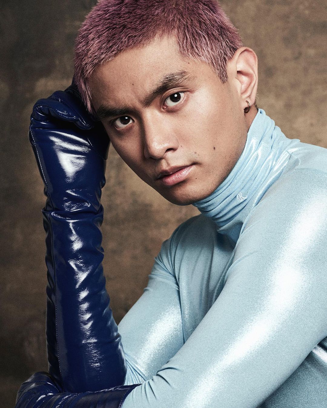 Portrait of a young adult male with light purple hair wearing a light blue turtleneck and dark blue, shiny gloves, indoor studio shot.