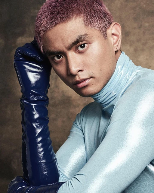 Portrait of a young adult male with light purple hair wearing a light blue turtleneck and dark blue, shiny gloves, indoor studio shot.